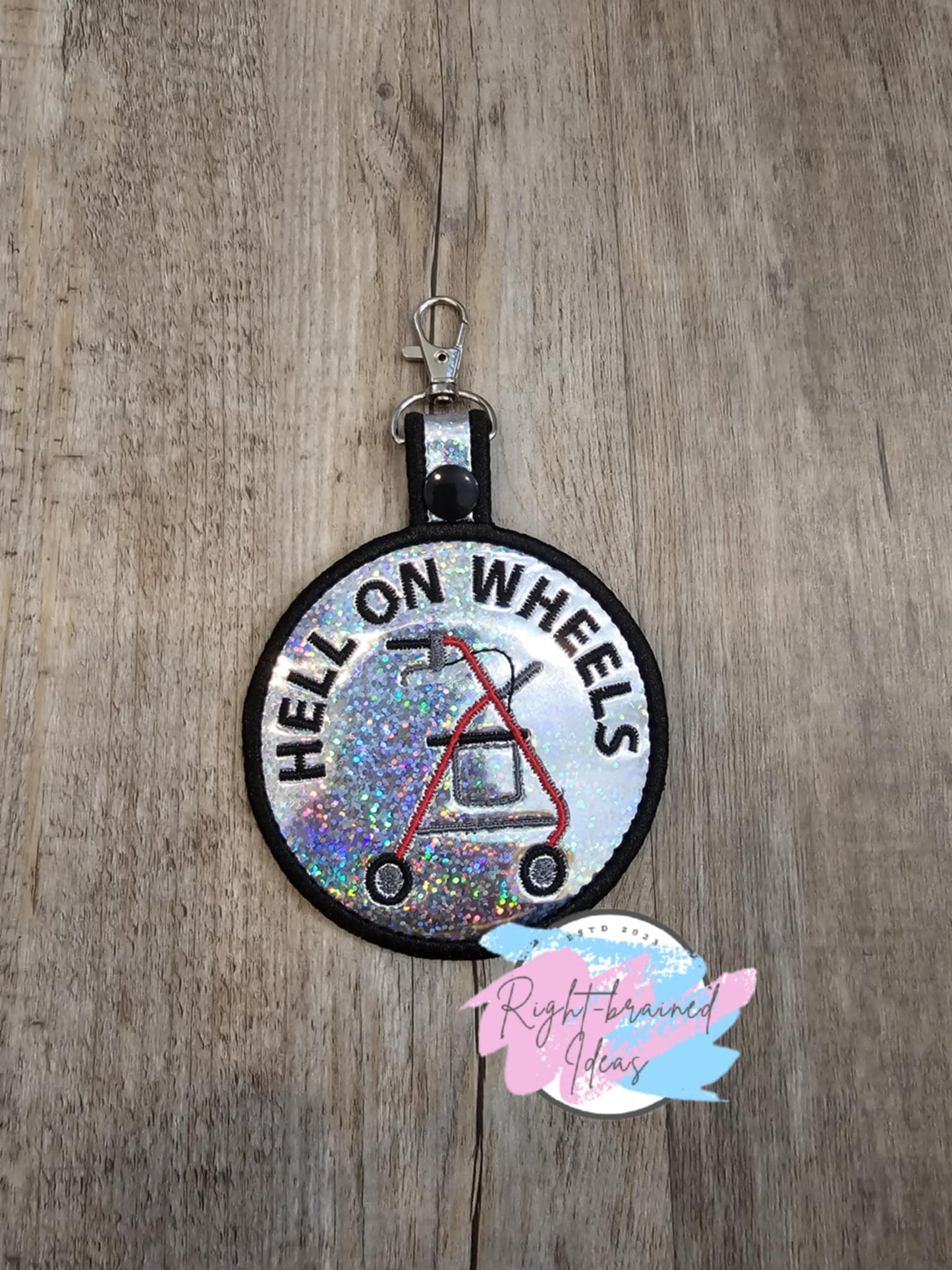 Rock and Roll Inspired Hell on Wheels Patch Tab. The tab is made with holographic sparkle silver vinyl and black, red, white and grey embroidery thread. It has a black plastic snap enclosing a metal d-ring clip.
Includes:
(1) 3.5in x 6in circular patch tab: "Hell on Wheels" around a rollator icon