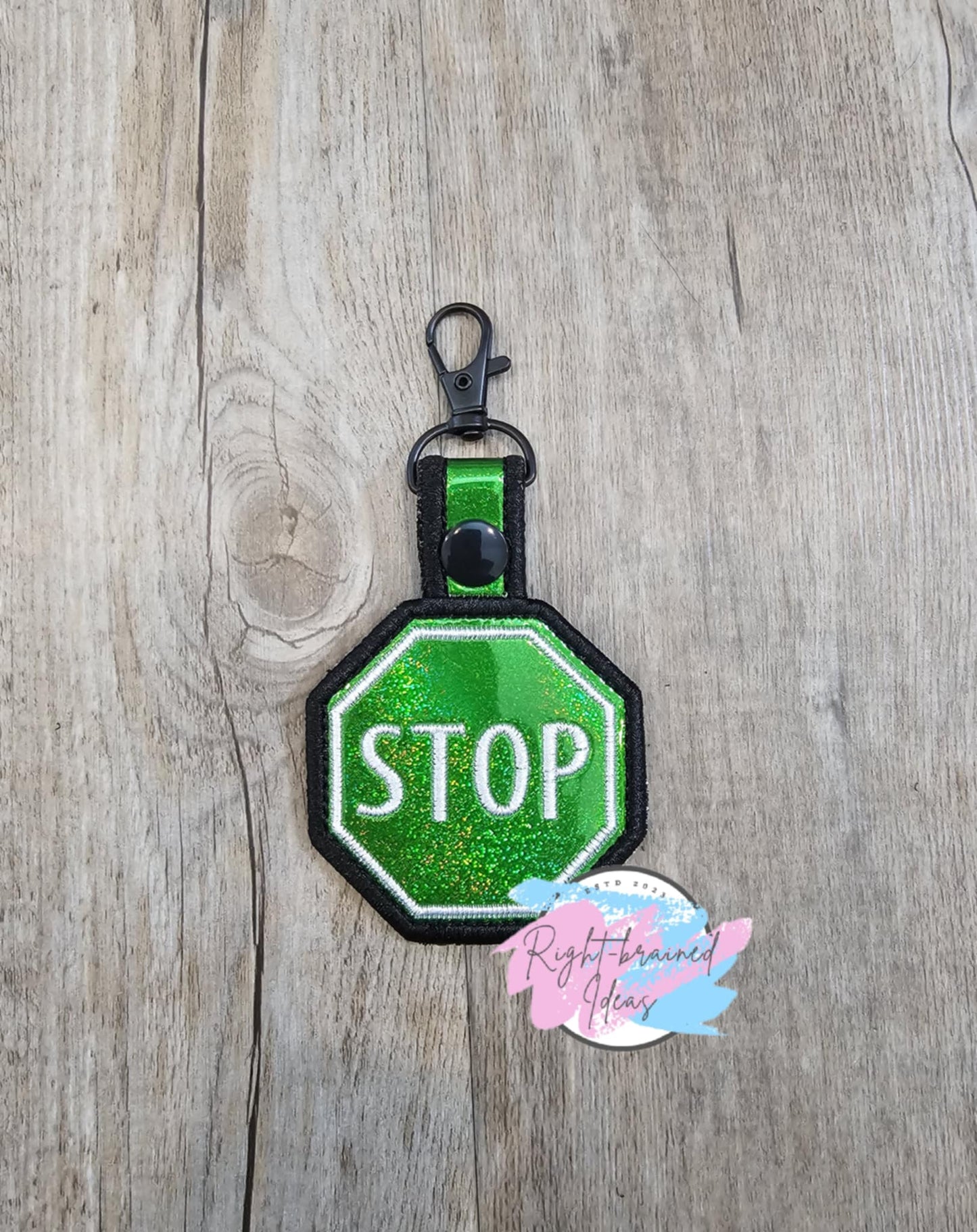 STOP sign Patch Tab. The tab is made with holographic sparkle green vinyl and black and white embroidery thread. It has a black plastic snap enclosing a metal d-ring clip.
(1) 2.5in hexagonal patch tab