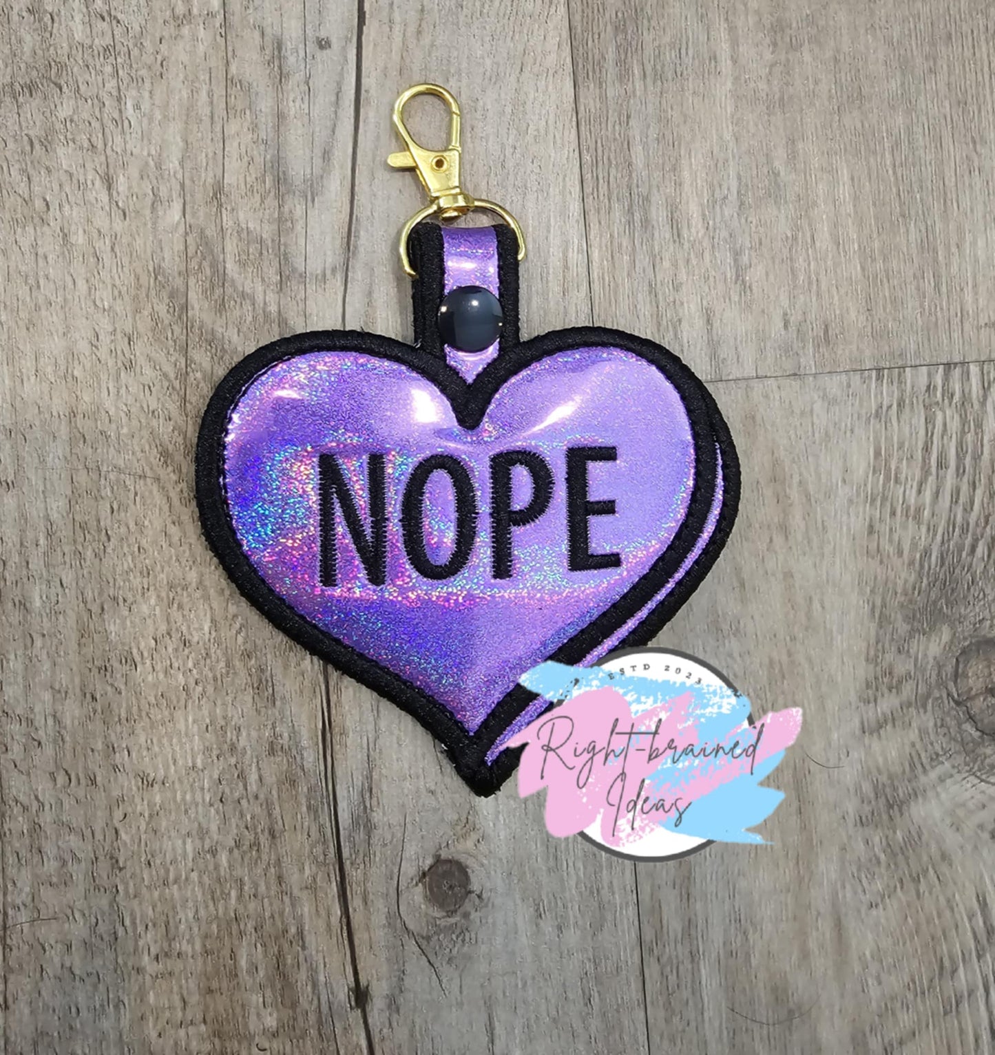 This is a Valentin's Day Conversation Heart inspired themed heart-shaped patch tab made with holographic light purple vinyl and black embroidery thread. The backside is made with black canvas fabric. It has a black plastic snap that encloses a gold metal d-ring clip.
(1) 4in x 6in heart-shaped patch tab