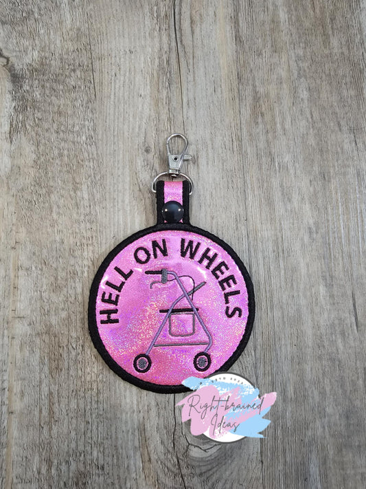 Rock and Roll Inspired Hell on Wheels Patch Tab. The tab is made with holographic sparkle pink vinyl and black, red, white and grey embroidery thread. It has a white plastic snap enclosing a metal d-ring clip.&nbsp;

Includes:
(1) 3.5in x 6in circular patch tab: "Hell on Wheels" around a rollator symbol