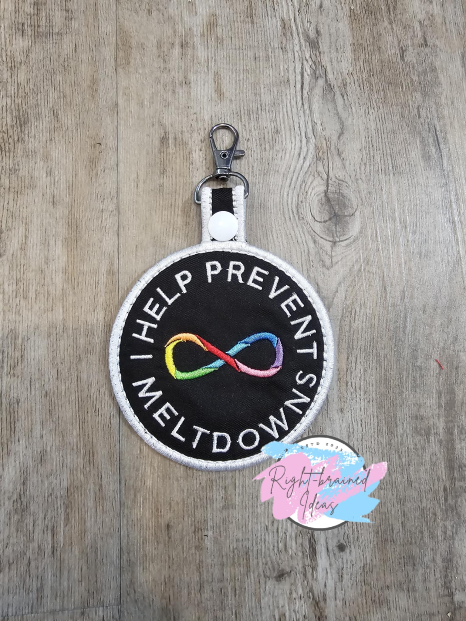 This is a Neurodivergent Inspired patch tab made of black twill with rainbow and white embroidery thread. It has a white plastic snap enclosing a metal d-ring clip.

(1) 3.5in x 6in circular patch tab: "Help Prevent Meltdowns" around a Rainbow Mobius/Infinity Loop in the center of the patch tab.